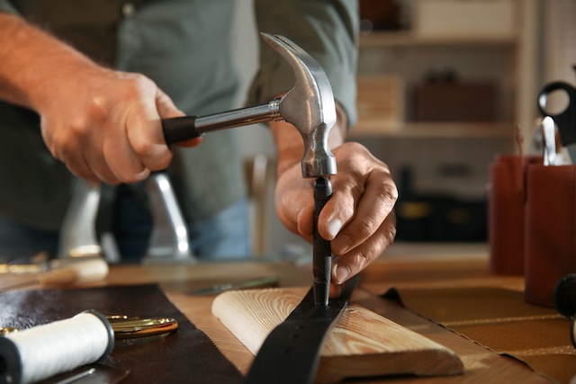 Close up of hands holding a hammer pounding a metal spike into a strip of leather on a work bench.