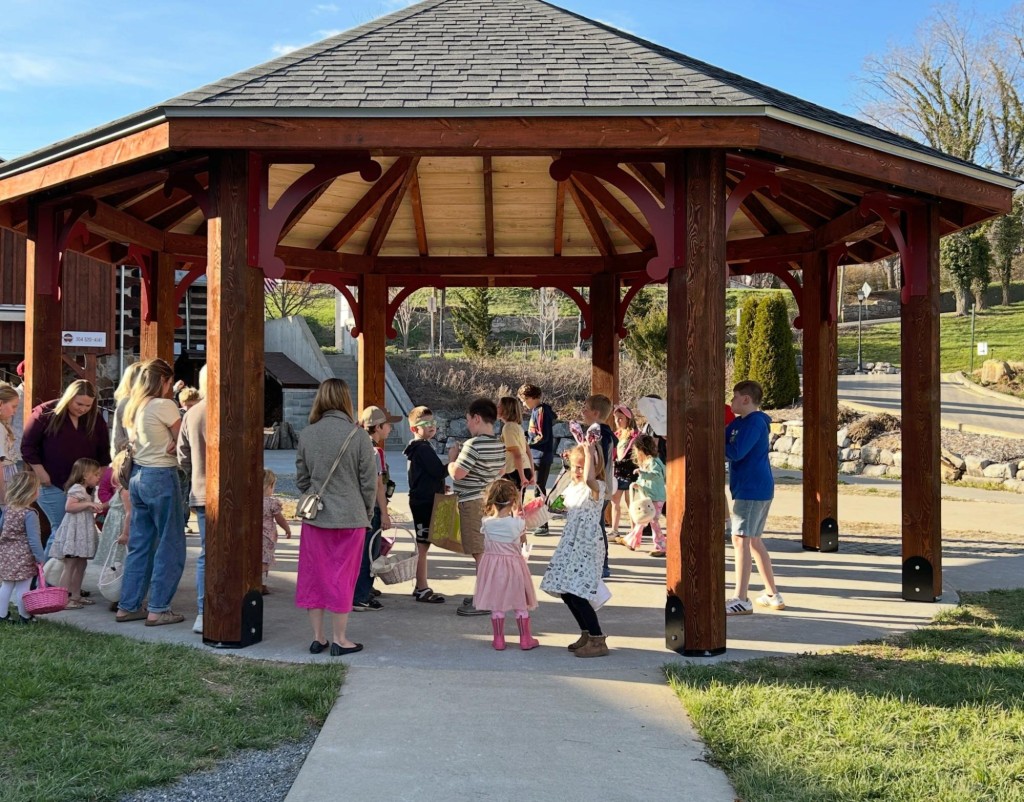 Children and parents stand under a wooden pavilion against a bright blue sky.