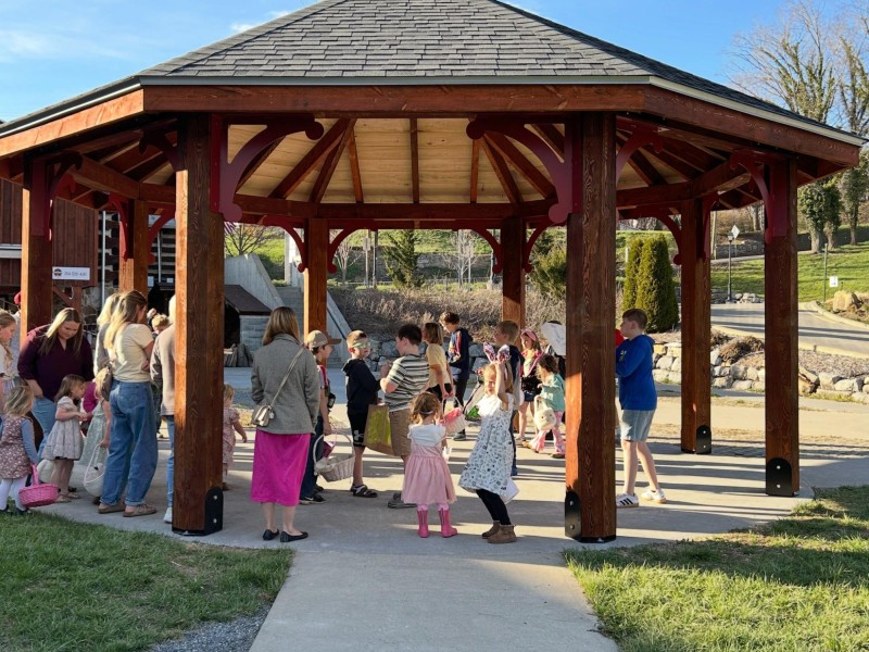 Children and parents stand under a wooden pavilion against a bright blue sky.
